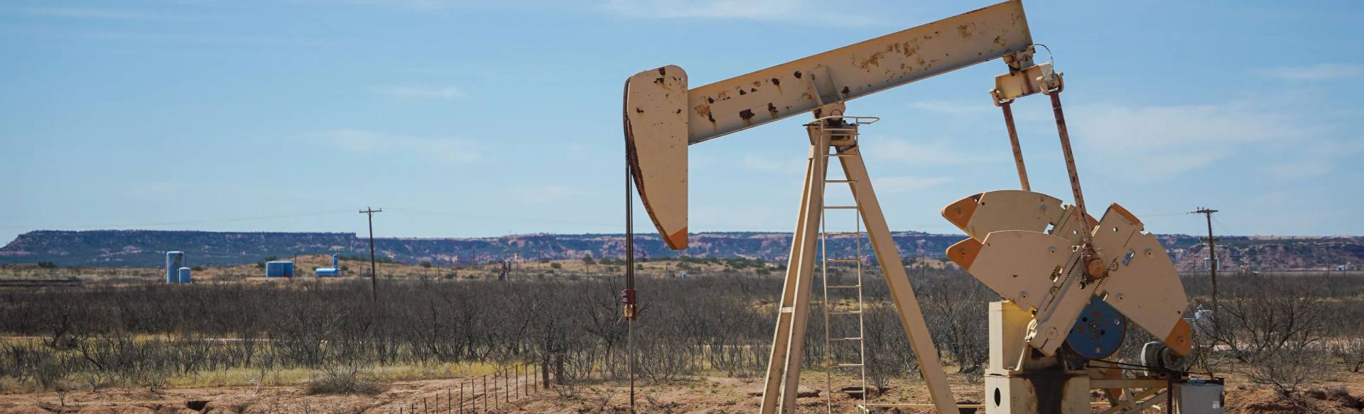 A working beam pump on a caliche pad in West Texas, mesquite scrub and tank batteries behind it under a clear sky