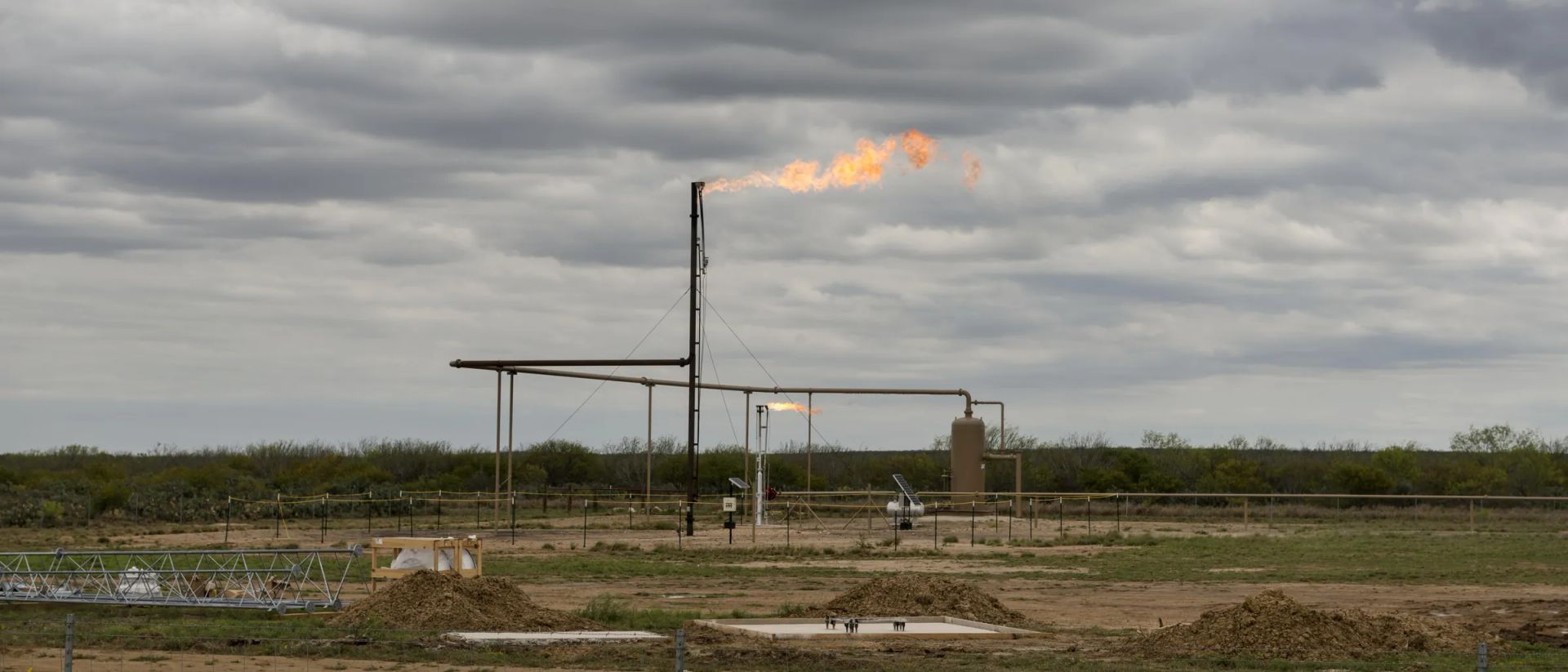 A lit flare burning off a stack over a fenced South Texas wellsite with a vertical separator, a horizontal vessel and an overhead pipe rack, under a heavy grey sky