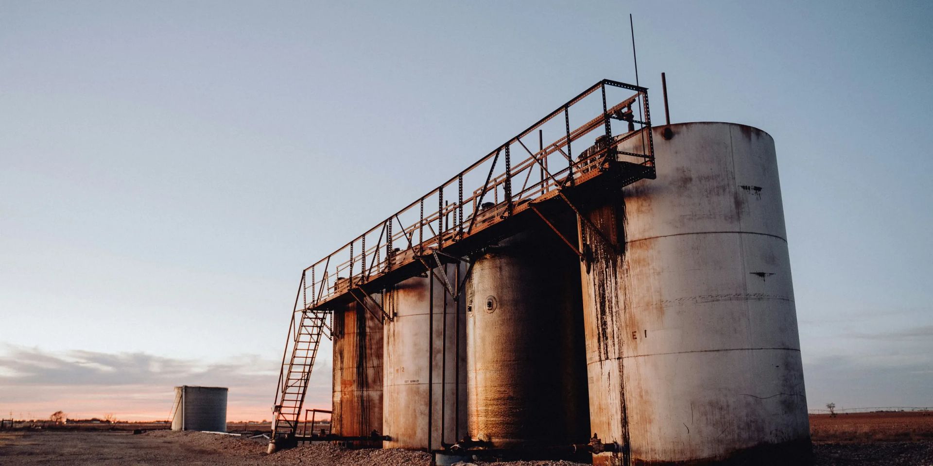 Four bolted steel stock tanks with a stair and catwalk on a gravel pad at dusk, with a smaller vessel at left and a flat farmed horizon