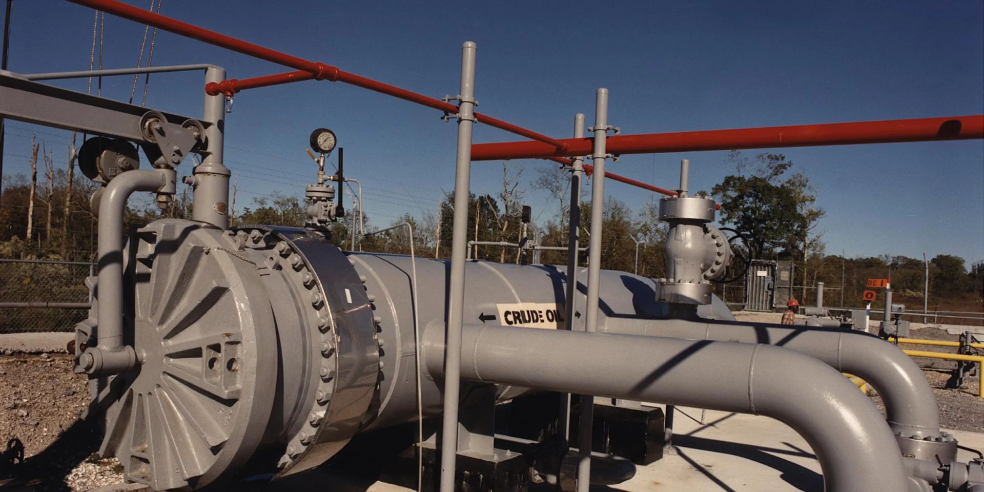 A pig launcher with a spoked quick-opening closure, barrel, eccentric reducer, kicker line and launcher valve on a concrete plinth, under a deep blue sky