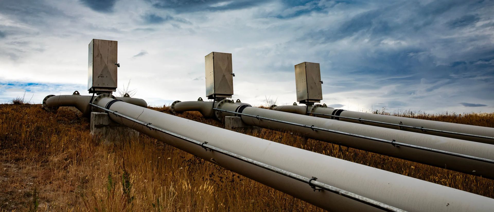 Three above-ground steel pipelines with flanged spools and stainless junction boxes, running across dry grass under a heavy cloud sky