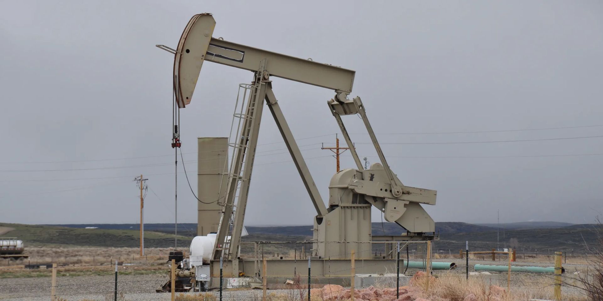 A beam pumping unit on a fenced gravel lease, with a coated pipe spool on the ground and low hills behind under an overcast sky