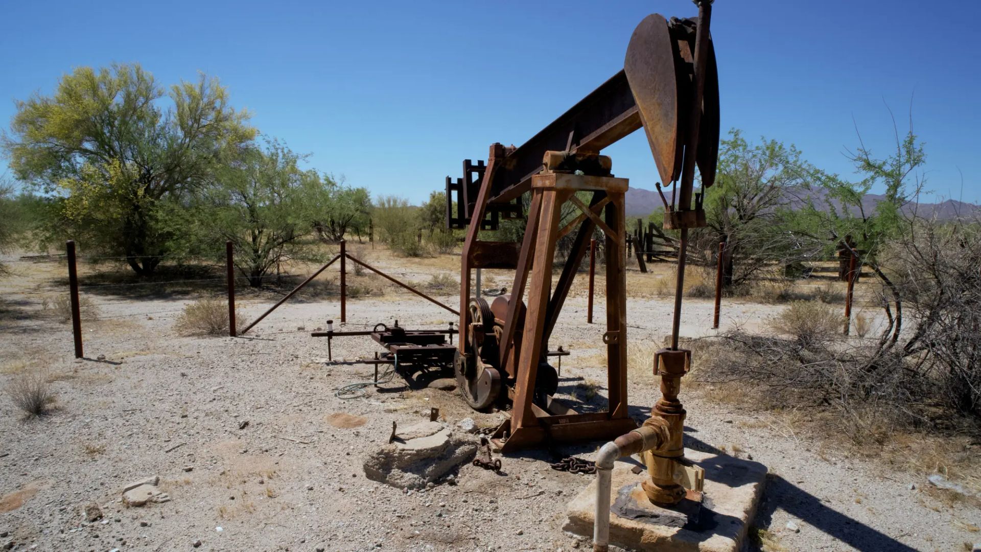 A weathered beam pumping unit and wellhead on a concrete pad, on a fenced desert lease with mesquite and hills behind