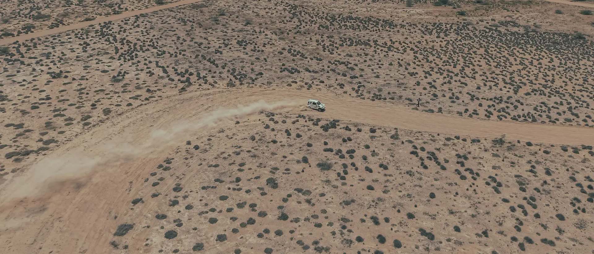 Drone view of a white four-by-four raising a dust trail along a curving dirt track through desert scrub
