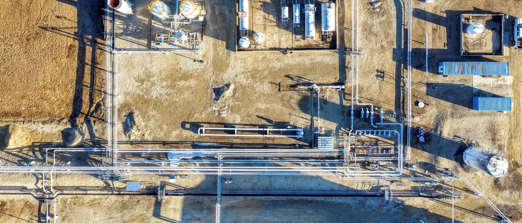 Straight-down aerial of a facility on gravel: pipe runs, valve manifolds, vessels, skid buildings and long winter shadows
