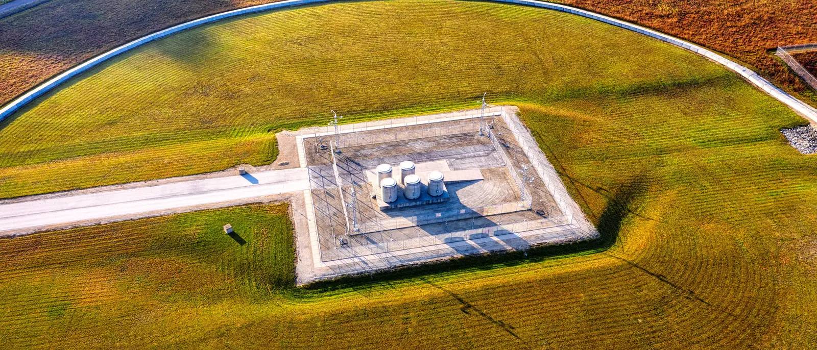 Drone view of a fenced concrete pad holding several capped vessels, with a single gravel access road running to it across a field