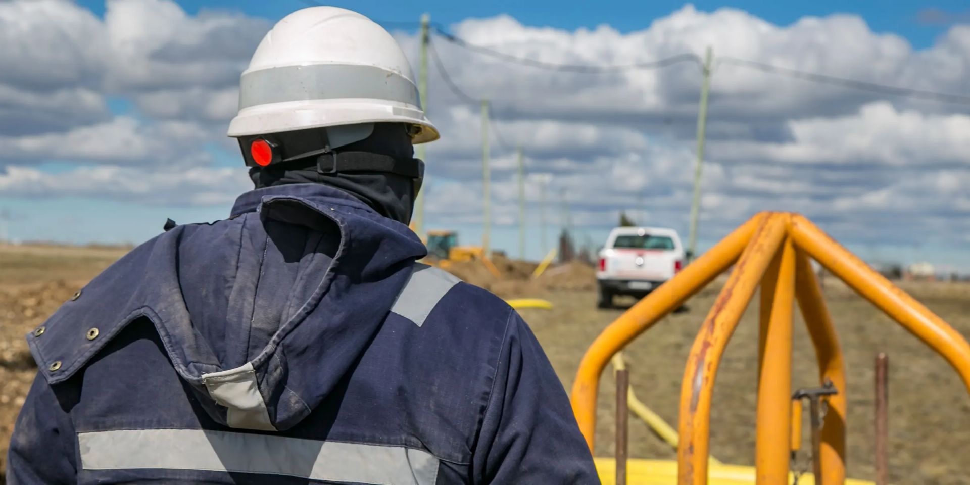 A field worker in a navy coverall with reflective tape and a white hard hat, seen from behind at a gas line tie-in, with yellow pipe, a pickup and an excavator beyond