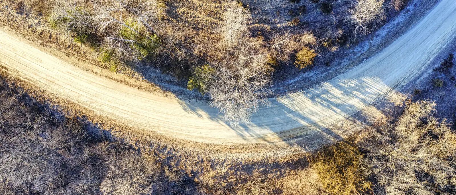 Straight-down aerial of a gravel road curving through winter scrub, with tyre tracks worn into the surface