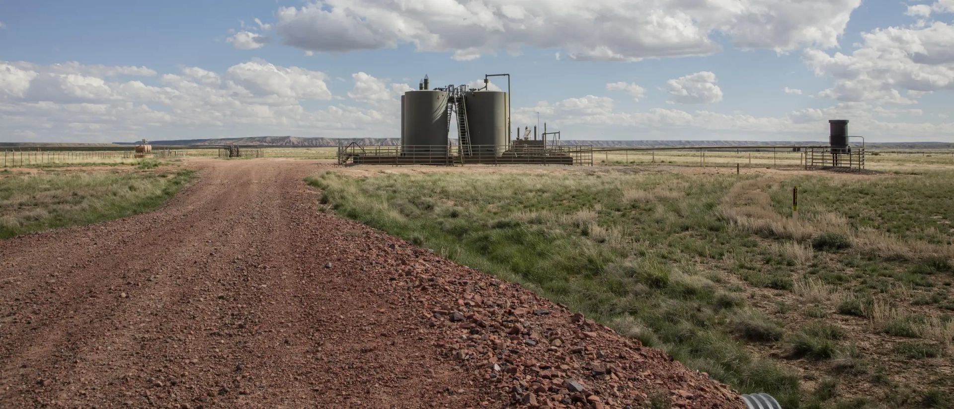 A red gravel lease road running straight to a fenced pad carrying two bolted stock tanks with a stair and catwalk, a separator and a heater, under a wide cumulus sky