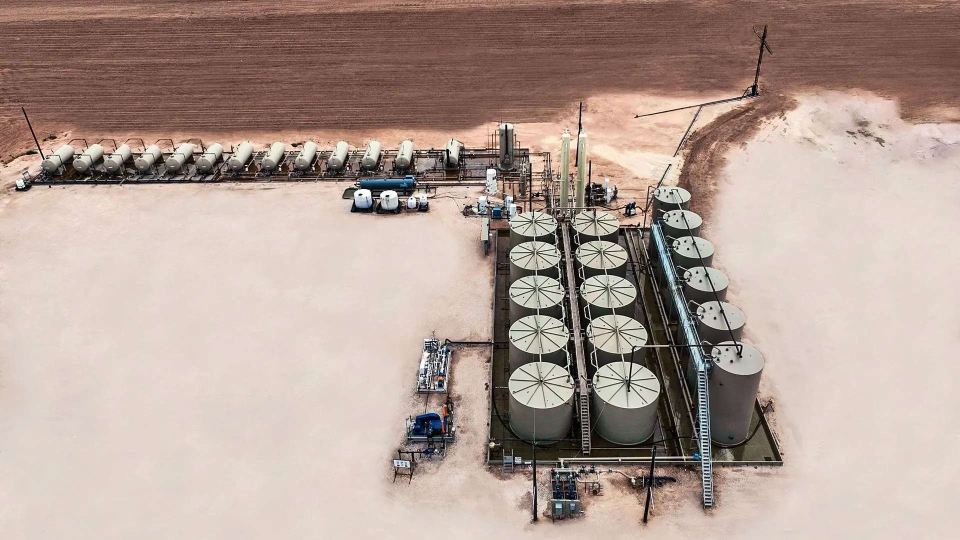 Aerial view of a production facility: a row of horizontal separators along the pad edge, vertical vessels, and a bank of stock tanks inside a lined containment with stairways and catwalks