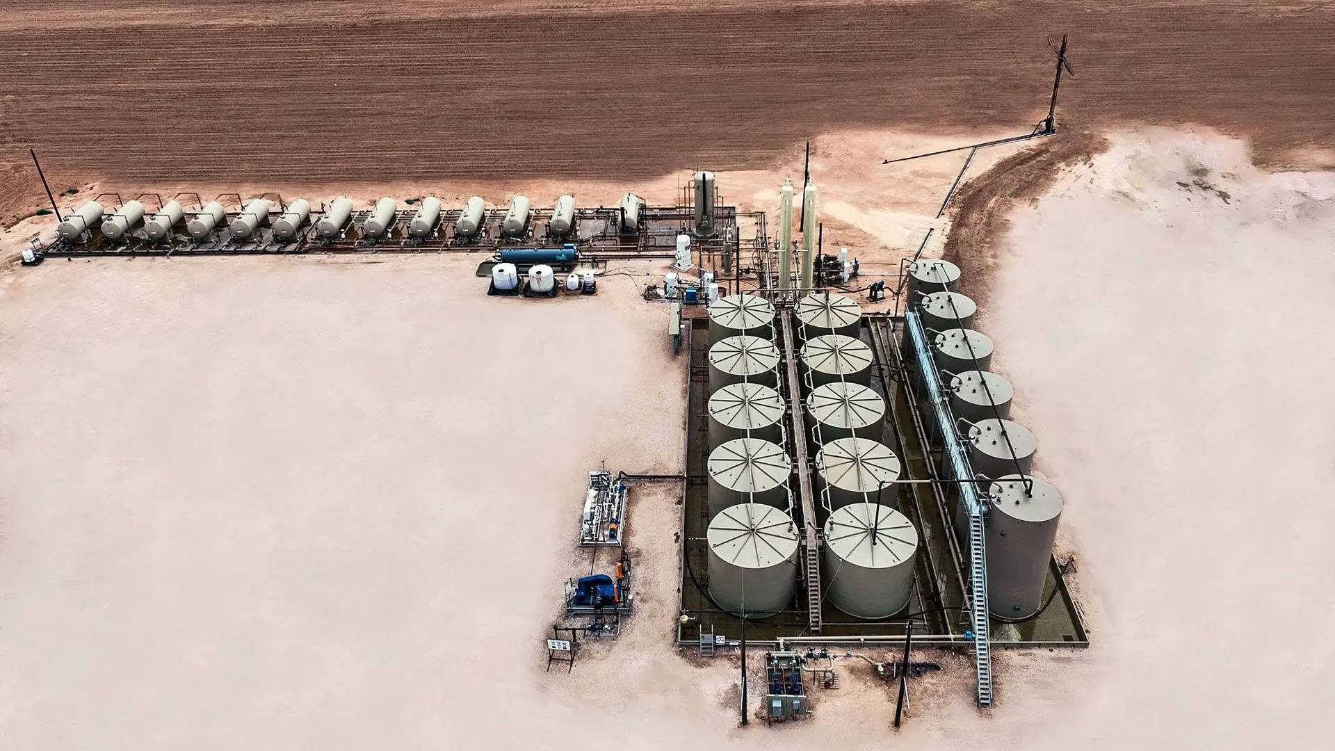 Aerial view of a production facility: a row of horizontal separators along the pad edge, vertical vessels, and a bank of stock tanks inside a lined containment with stairways and catwalks