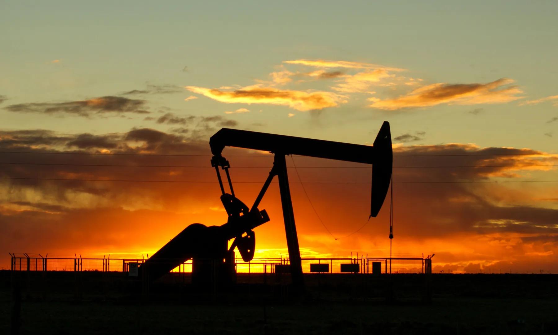 A pumping unit in silhouette against an orange sky at first light, a fenced wellsite and power lines on the flat horizon