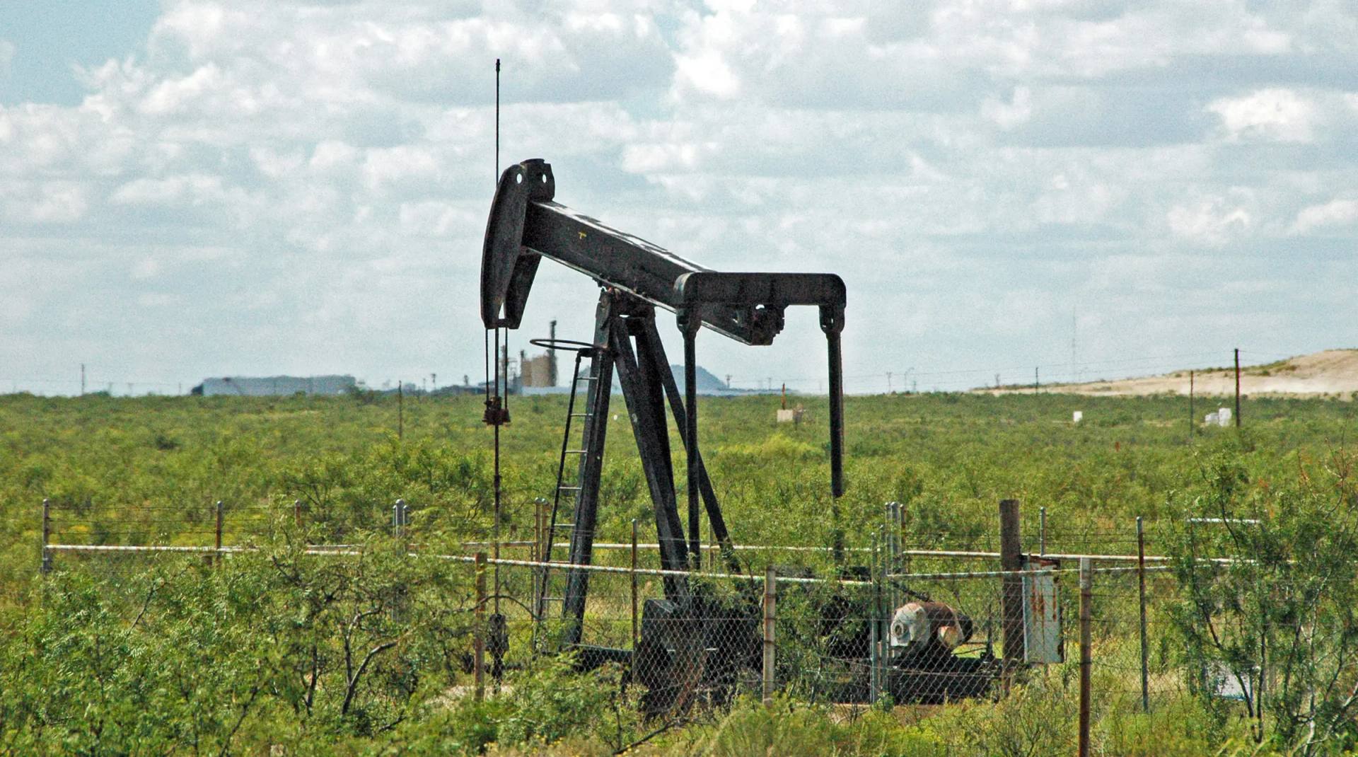 A black pumping unit in mesquite scrub beside its fenced wellhead