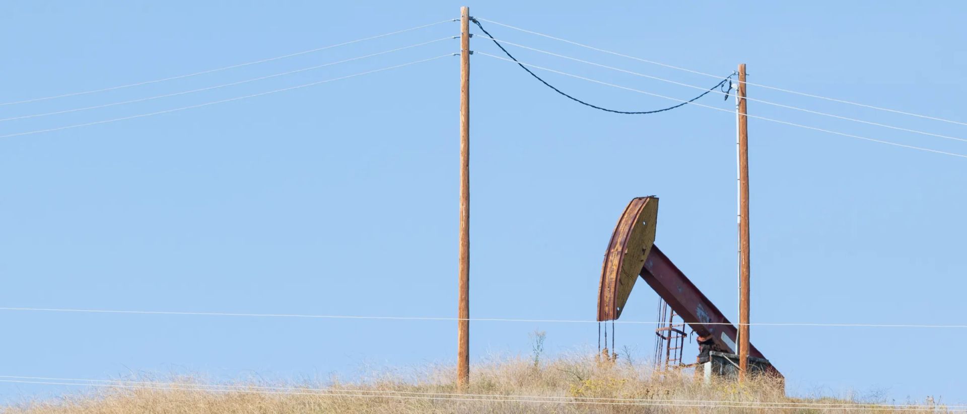 A single pumping unit on a grassy ridge between two wooden power poles, under an open blue sky