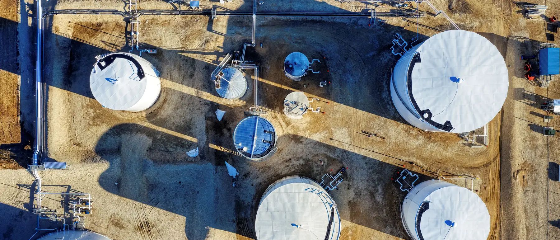 Storage tanks, small process vessels and pipe racks on a gravel pad, photographed straight down from a drone in low winter sun