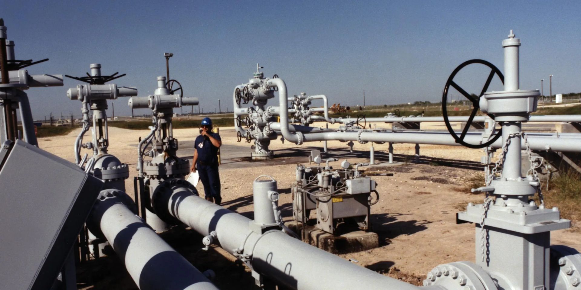 A line of wellheads with gate valves, handwheels and gear operators on a sand pad, with two painted flowlines running to the foreground