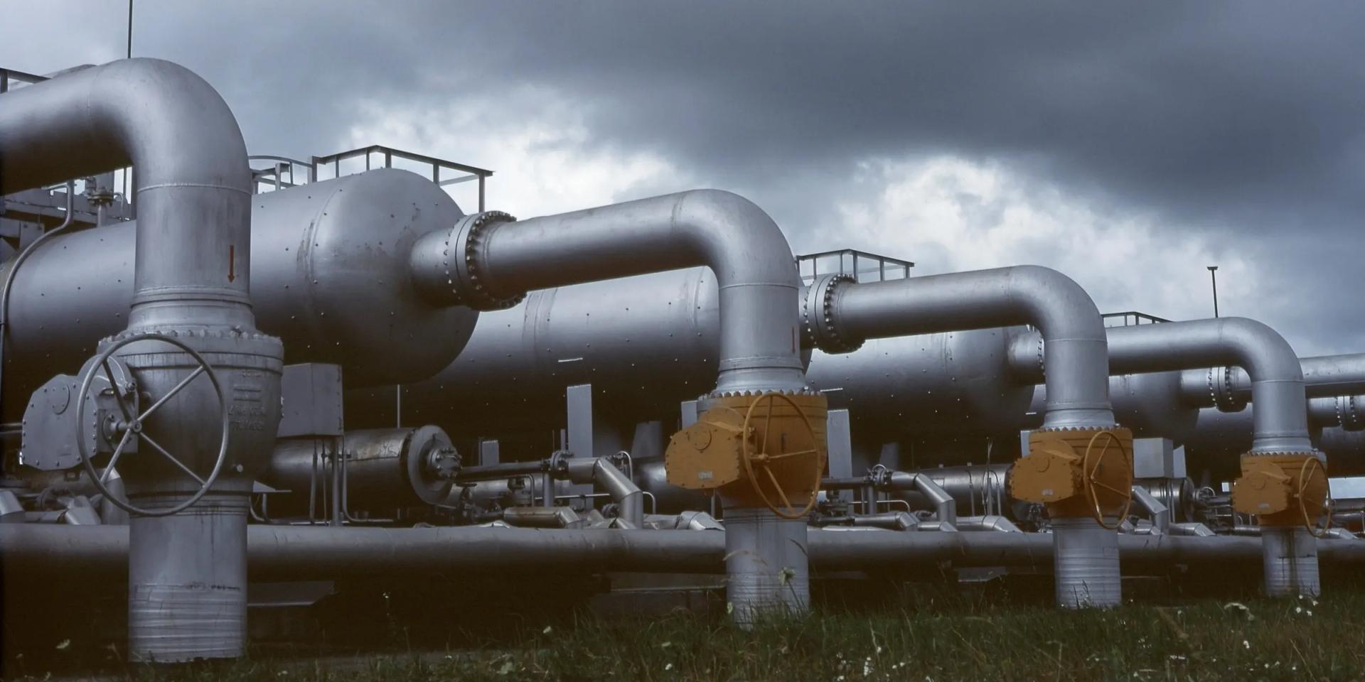 A gas facility valve manifold under a heavy sky: a hand-wheel gate valve, three yellow actuators and process vessels behind them