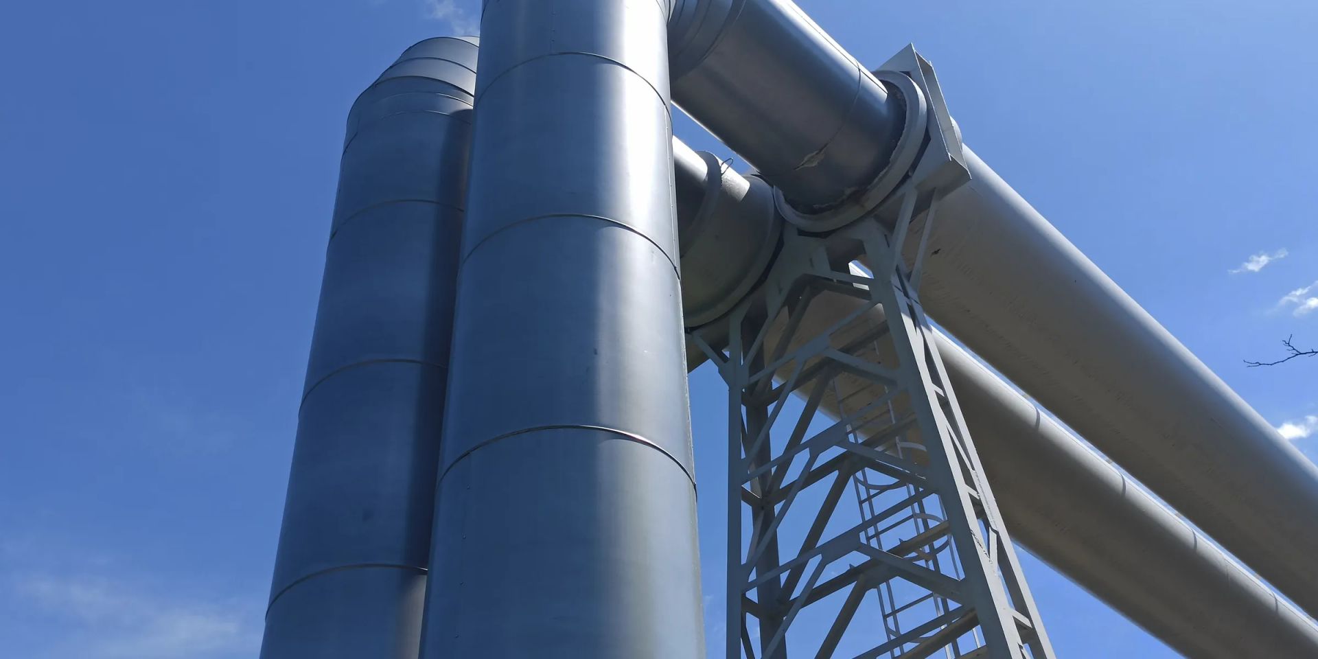 Insulated steel process pipes turning across a braced lattice support tower against a clear blue sky
