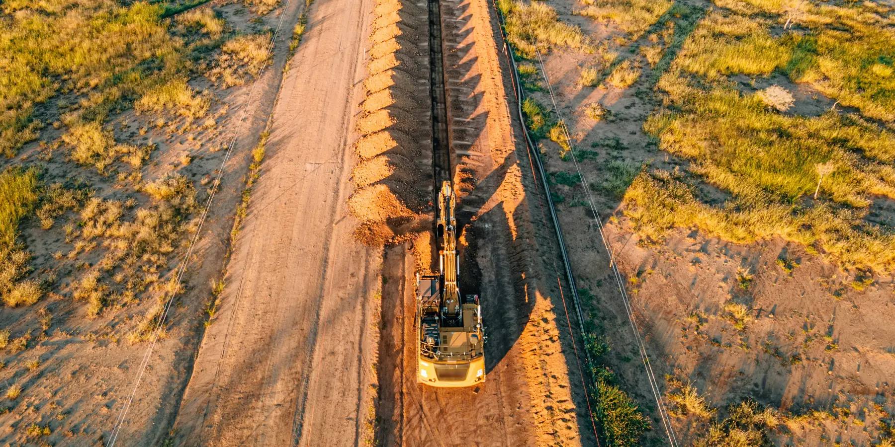 Looking straight down a new pipeline right of way at golden hour: an open trench with spoil along it, pipe strung beside the ditch line, a tracked excavator working and a parallel haul road through rangeland