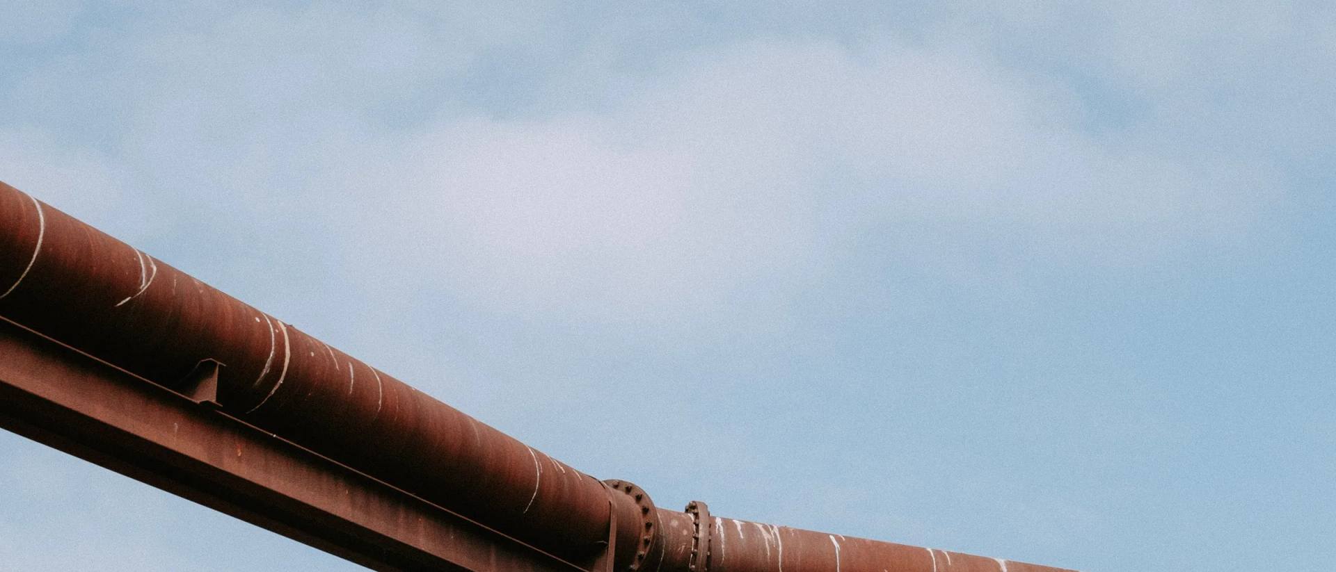 A weathered steel pipeline on a braced trestle, running across a pale sky above open grass