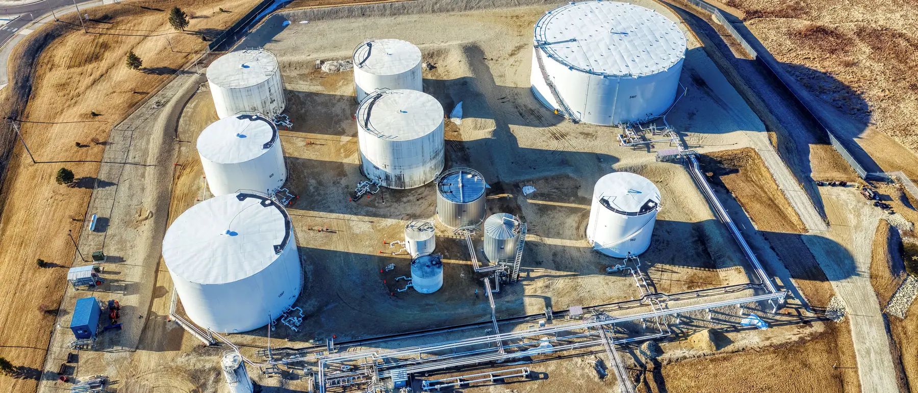 Aerial view of a fenced gravel yard with white upright storage tanks, smaller stainless vessels, pipe racks and blue skid buildings, with brown winter fields beyond